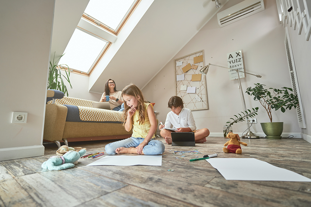 Cute excited little girl sitting on the floor, drawing with colorful pencils while her brother doing homework online, happy mother sitting on sofa, drinking coffee and using laptop ductless AC in attic office
